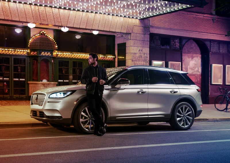A 2022 Lincoln Corsair SUV is parked outside a theater as the driver relaxes against the frame and lights illuminate the floating roofline and body | Pugmire Lincoln of Marietta in Marietta GA