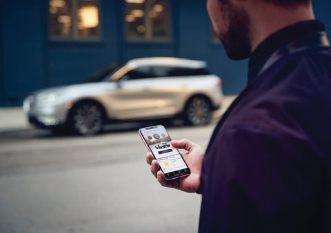A man is holding his phone looking at the Lincoln Way™ App and we see his 2022 Lincoln Corsair SUV in the background against a blue brick wall | Pugmire Lincoln of Marietta in Marietta GA